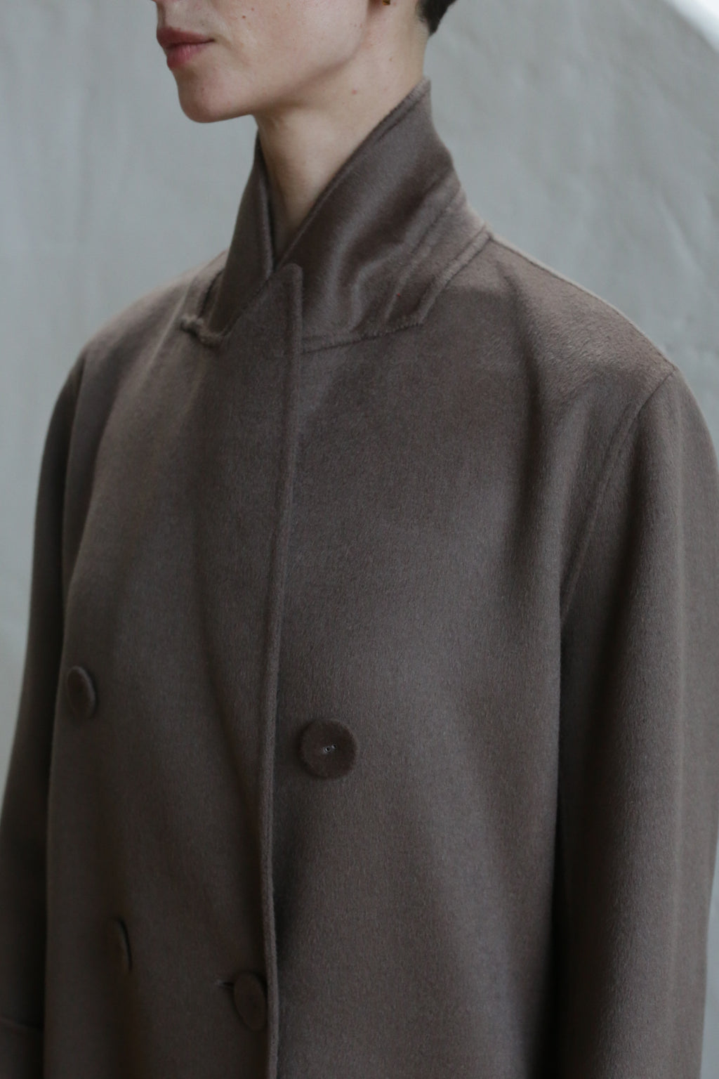 A person wearing the Doublebreasted Coat in taupe, featuring a high collar and large buttons, is shown from shoulders to chin against a neutral background.