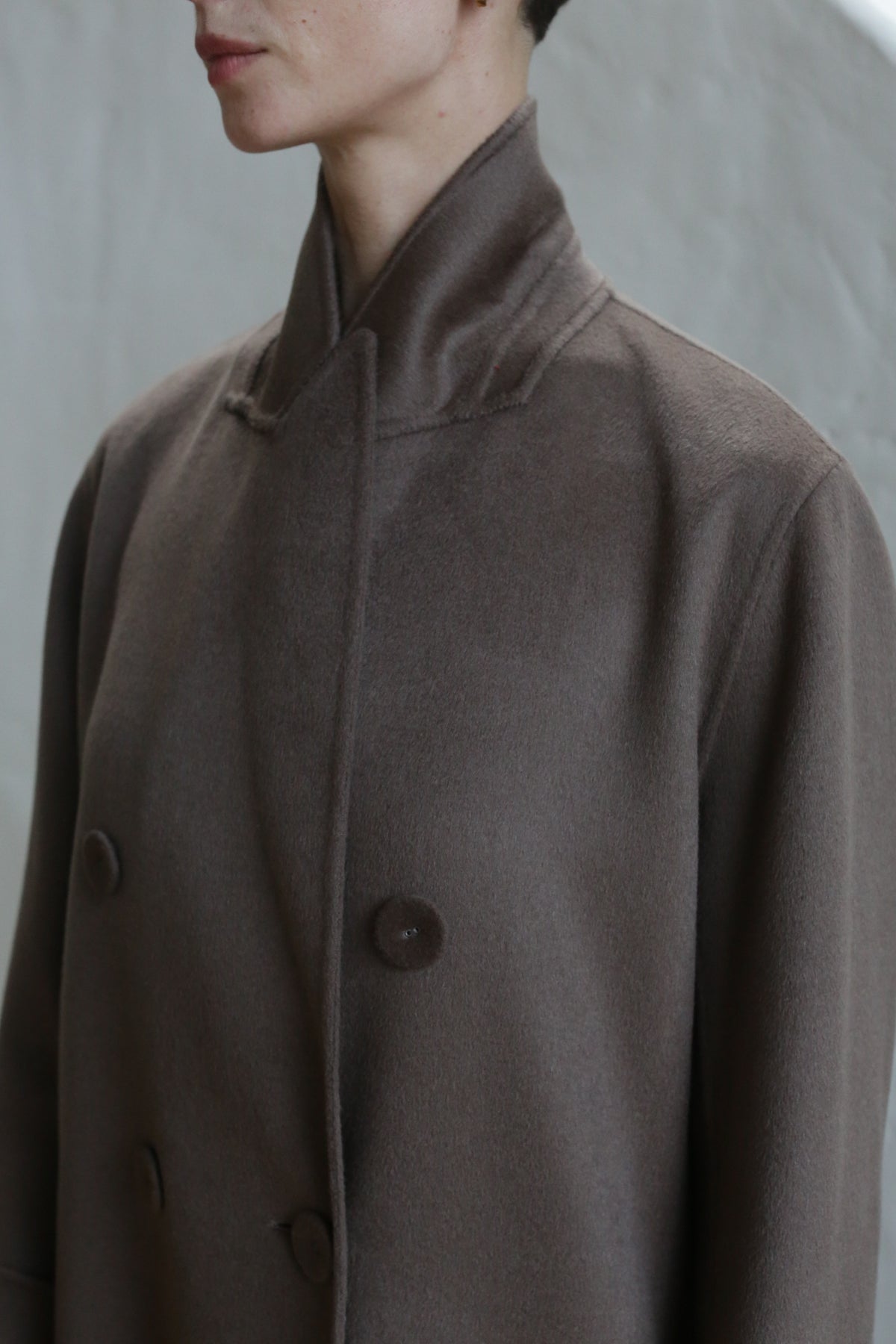 A person wearing the Doublebreasted Coat in taupe, featuring a high collar and large buttons, is shown from shoulders to chin against a neutral background.