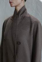 Load image into Gallery viewer, A person wearing the Doublebreasted Coat in taupe, featuring a high collar and large buttons, is shown from shoulders to chin against a neutral background.