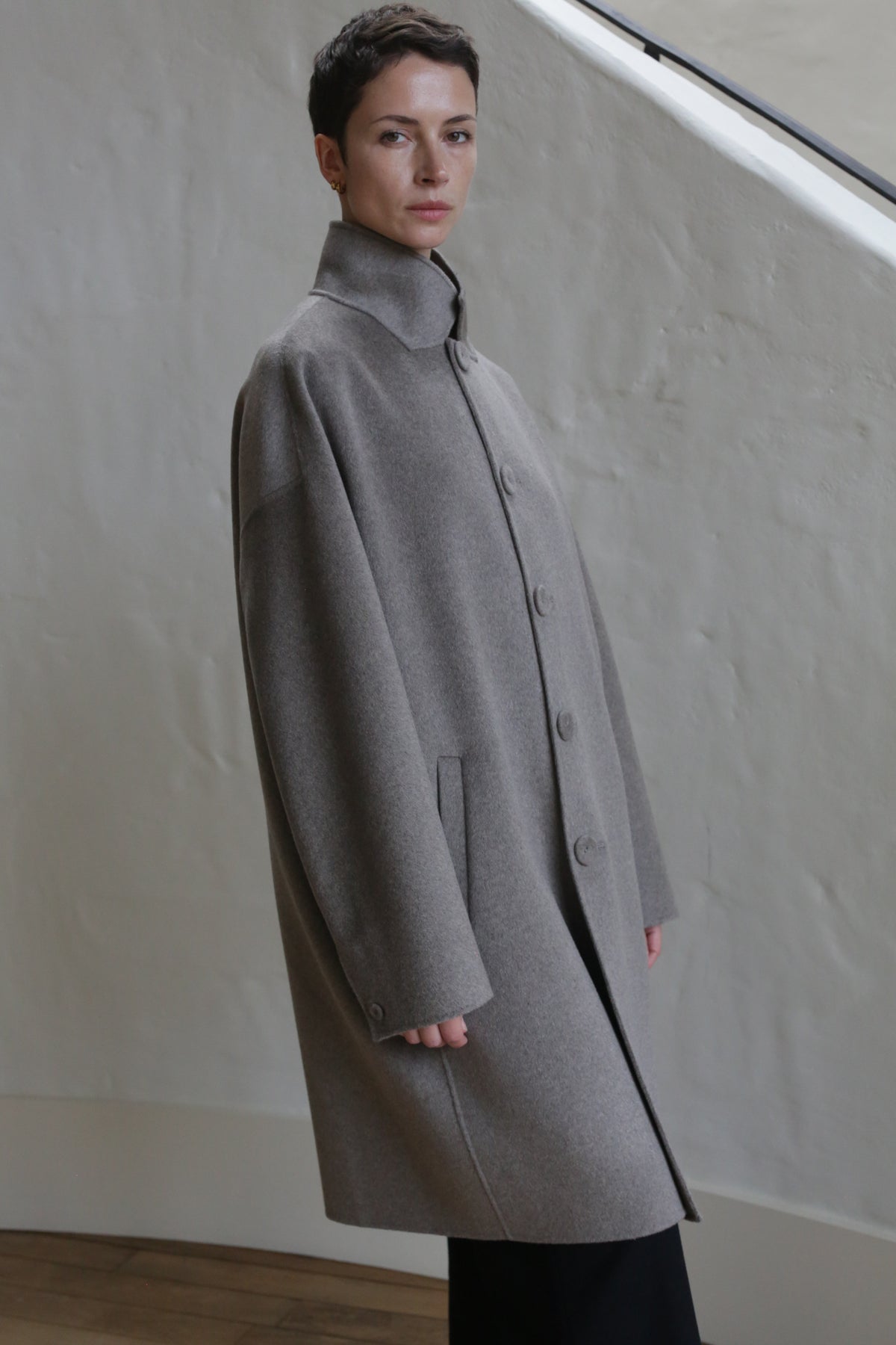 A man with short dark hair wears the Men's Coat | Undyed Natural, a loose-fitting, light-gray wool coat with large buttons, standing indoors by a textured beige wall and staircase railing.