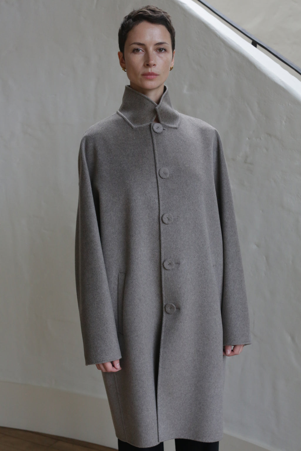 A person with short hair wears the Men's Coat in Undyed Natural and stands indoors before a light-colored wall and staircase.
