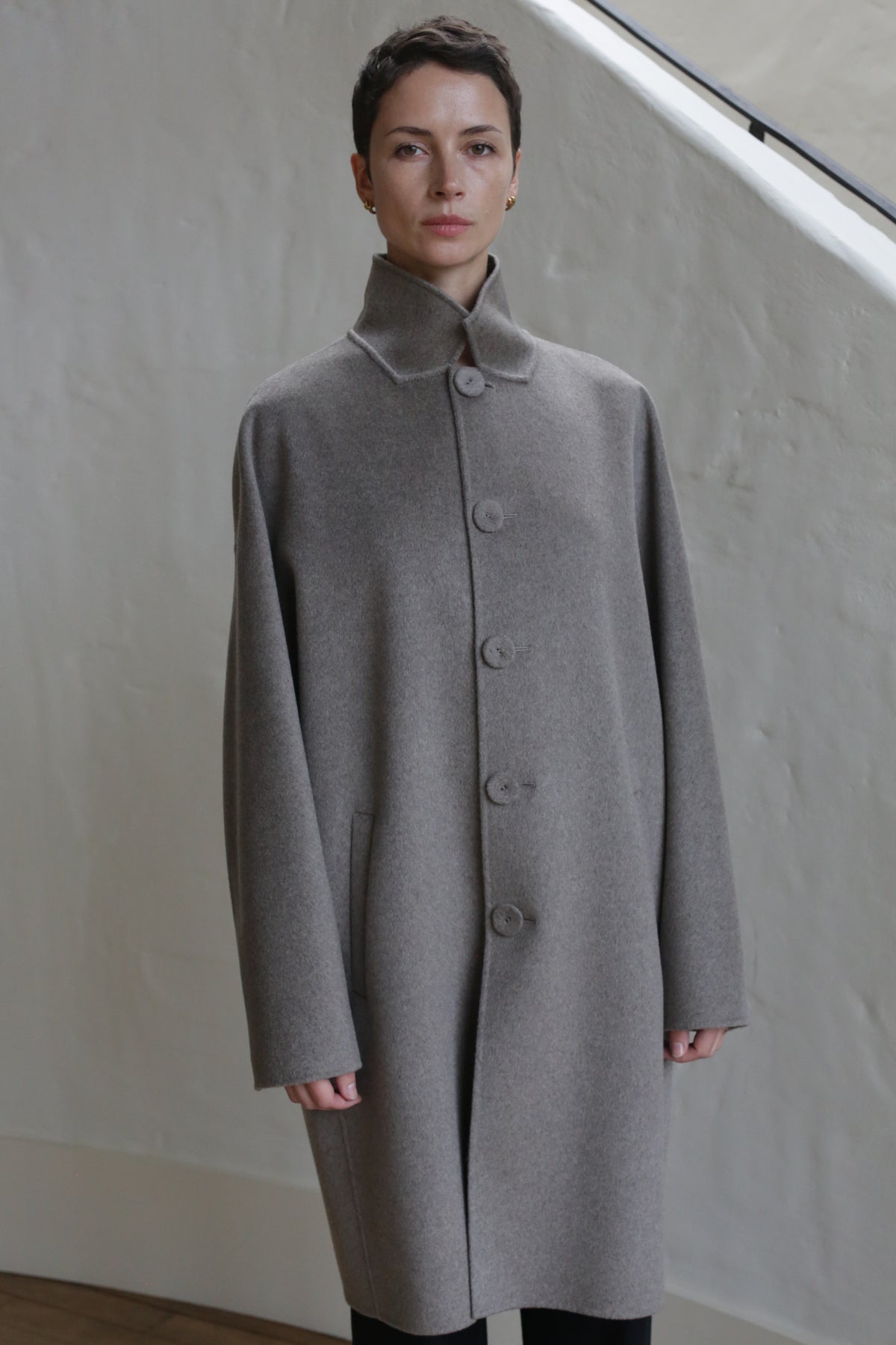 A person with short hair wears the Men's Coat in Undyed Natural and stands indoors before a light-colored wall and staircase.