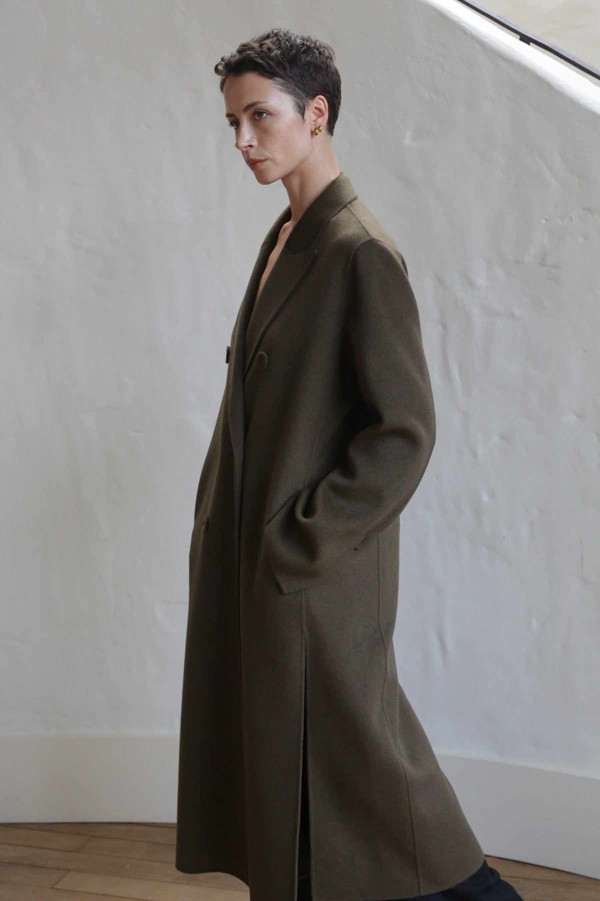 A person with short hair stands indoors against a white textured wall, wearing the Doublebreasted Coat | Olive with hands in the pockets and gazing thoughtfully to the side.