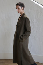 Load image into Gallery viewer, A person with short hair stands indoors against a white textured wall, wearing the Doublebreasted Coat | Olive with hands in the pockets and gazing thoughtfully to the side.