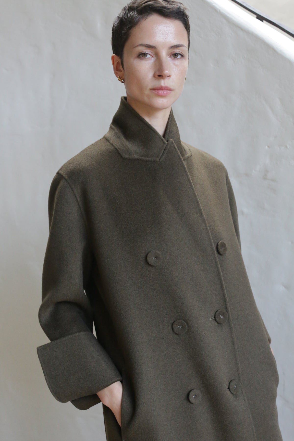 A person with short dark hair wears the Doublebreasted Coat | Olive, featuring wide sleeves, while standing against a light textured wall and looking at the camera with a neutral expression.