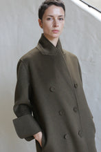 Load image into Gallery viewer, A person with short dark hair wears the Doublebreasted Coat | Olive, featuring wide sleeves, while standing against a light textured wall and looking at the camera with a neutral expression.