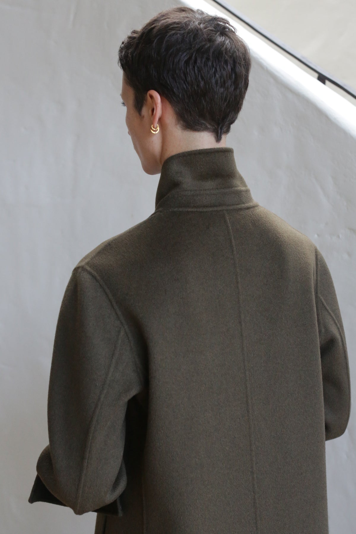 A person with short dark hair, wearing a gold earring and the Doublebreasted Coat | Olive, stands with their back to the camera against a light-colored background.
