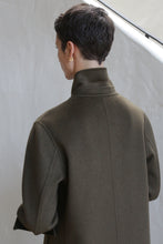 Load image into Gallery viewer, A person with short dark hair, wearing a gold earring and the Doublebreasted Coat | Olive, stands with their back to the camera against a light-colored background.