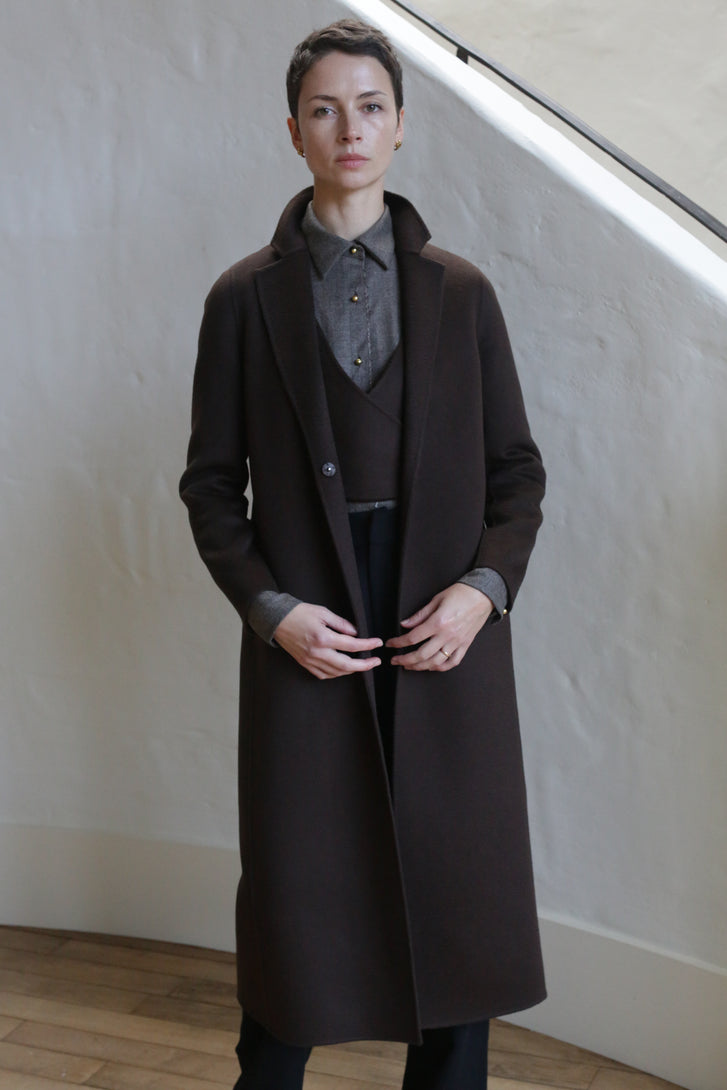 A person with short hair wears the X-Scarf Coat in Watermark Brown over a gray collared shirt and dark pants, standing indoors against a textured light-colored wall.