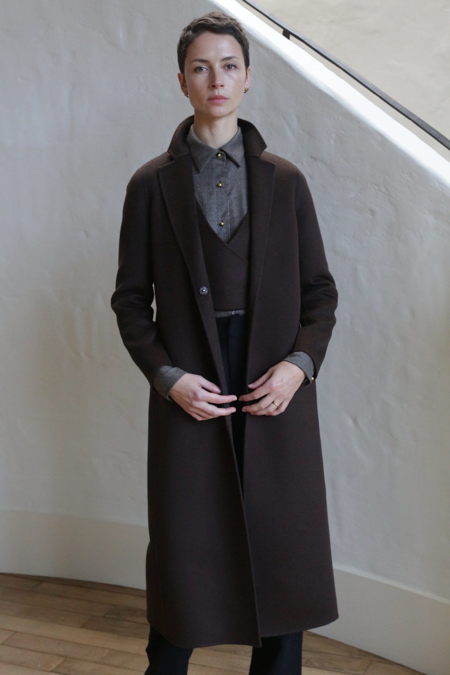 A person with short hair wears the X-Scarf Coat in Watermark Brown over a gray collared shirt and dark pants, standing indoors against a textured light-colored wall.