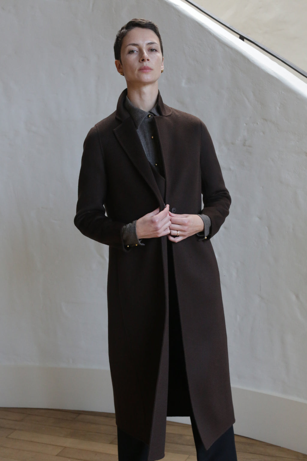 Wearing the X-Scarf Coat in Watermark Brown, a person with short dark hair stands indoors against a white textured wall and wooden floor, confidently facing the camera.