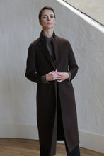 Load image into Gallery viewer, Wearing the X-Scarf Coat in Watermark Brown, a person with short dark hair stands indoors against a white textured wall and wooden floor, confidently facing the camera.
