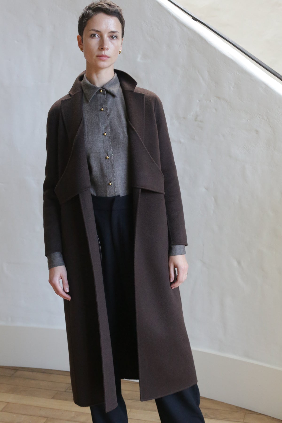 A person with short hair stands indoors in front of a white textured wall and staircase, wearing the X-Scarf Coat in Watermark Brown over a gray shirt and black pants.