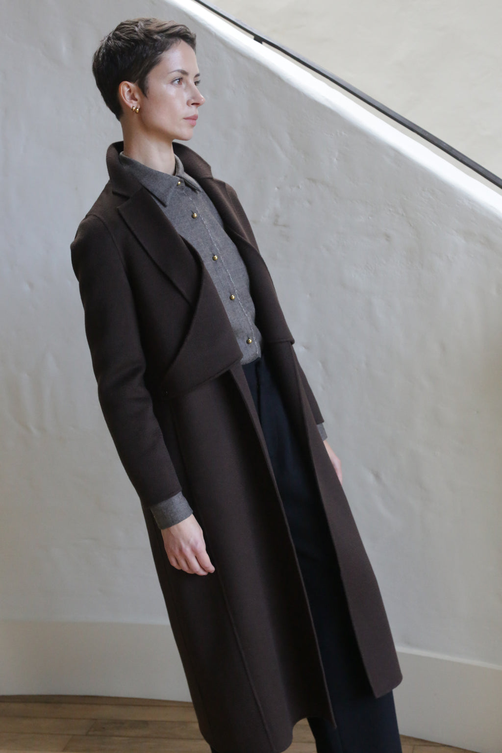 A person with short hair wearing the X-Scarf Coat in Watermark Brown over a gray button-up and black pants stands indoors against a light, textured wall, gazing thoughtfully to the side.