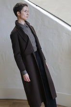 Load image into Gallery viewer, A person with short hair wearing the X-Scarf Coat in Watermark Brown over a gray button-up and black pants stands indoors against a light, textured wall, gazing thoughtfully to the side.