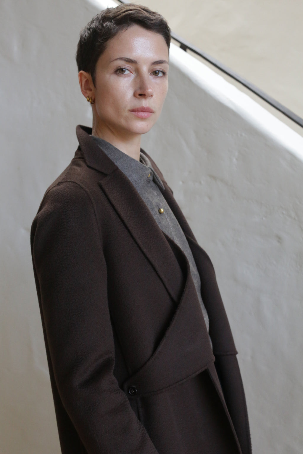 A person with short dark hair wears the X-Scarf Coat in Watermark Brown over a gray button-up shirt, standing indoors against a textured light wall and looking calmly at the camera with a neutral expression.