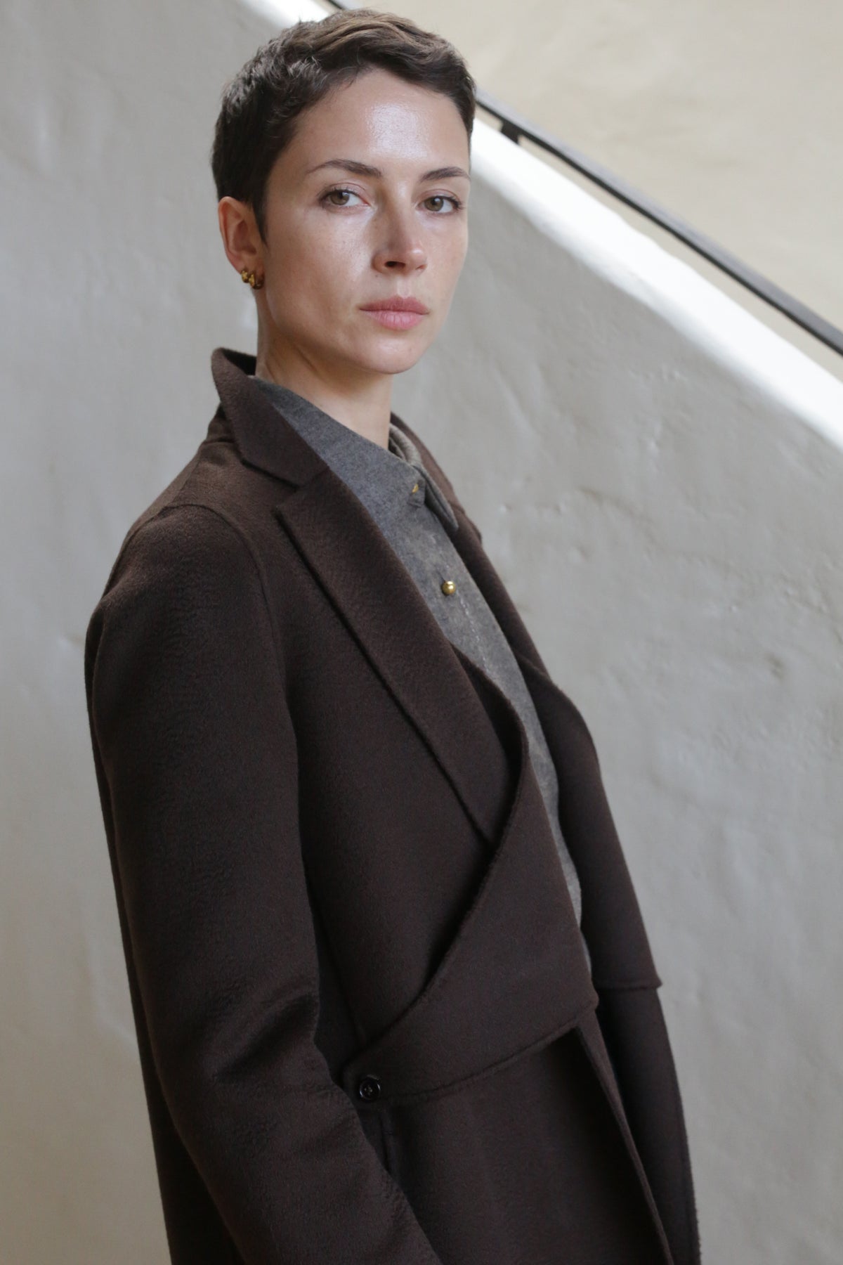 A person with short dark hair wears the X-Scarf Coat in Watermark Brown over a gray button-up shirt, standing indoors against a textured light wall and looking calmly at the camera with a neutral expression.