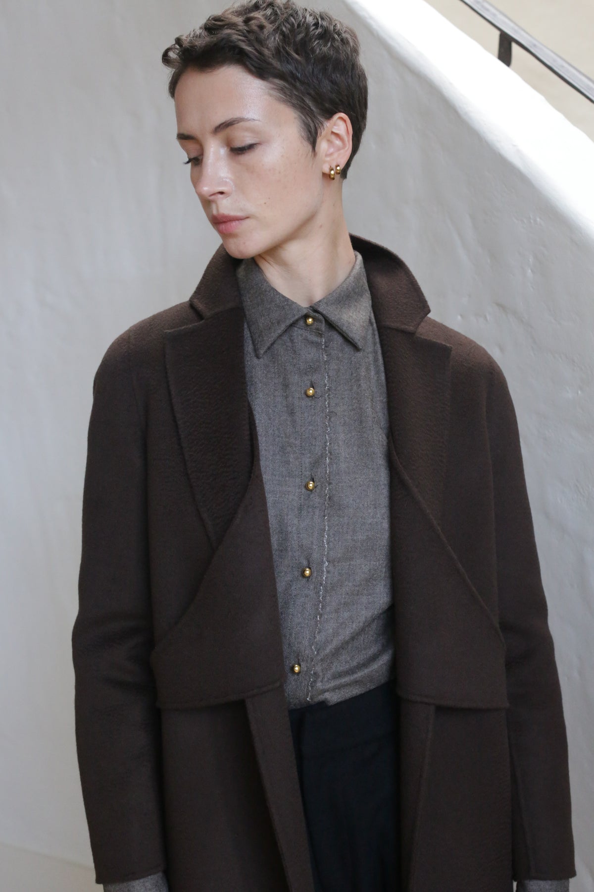 A person with short hair, wearing the X-Scarf Coat in Watermark Brown over a buttoned gray shirt, stands indoors against a textured white wall, looking down and to the side.