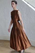 Load image into Gallery viewer, A person stands indoors on a wooden floor against a textured white wall, wearing the Hand Pleated Column Dress in copper silk charmeuse. This sleeveless dress features a fitted bodice, mid-length flowing skirt, and elegant pleats.