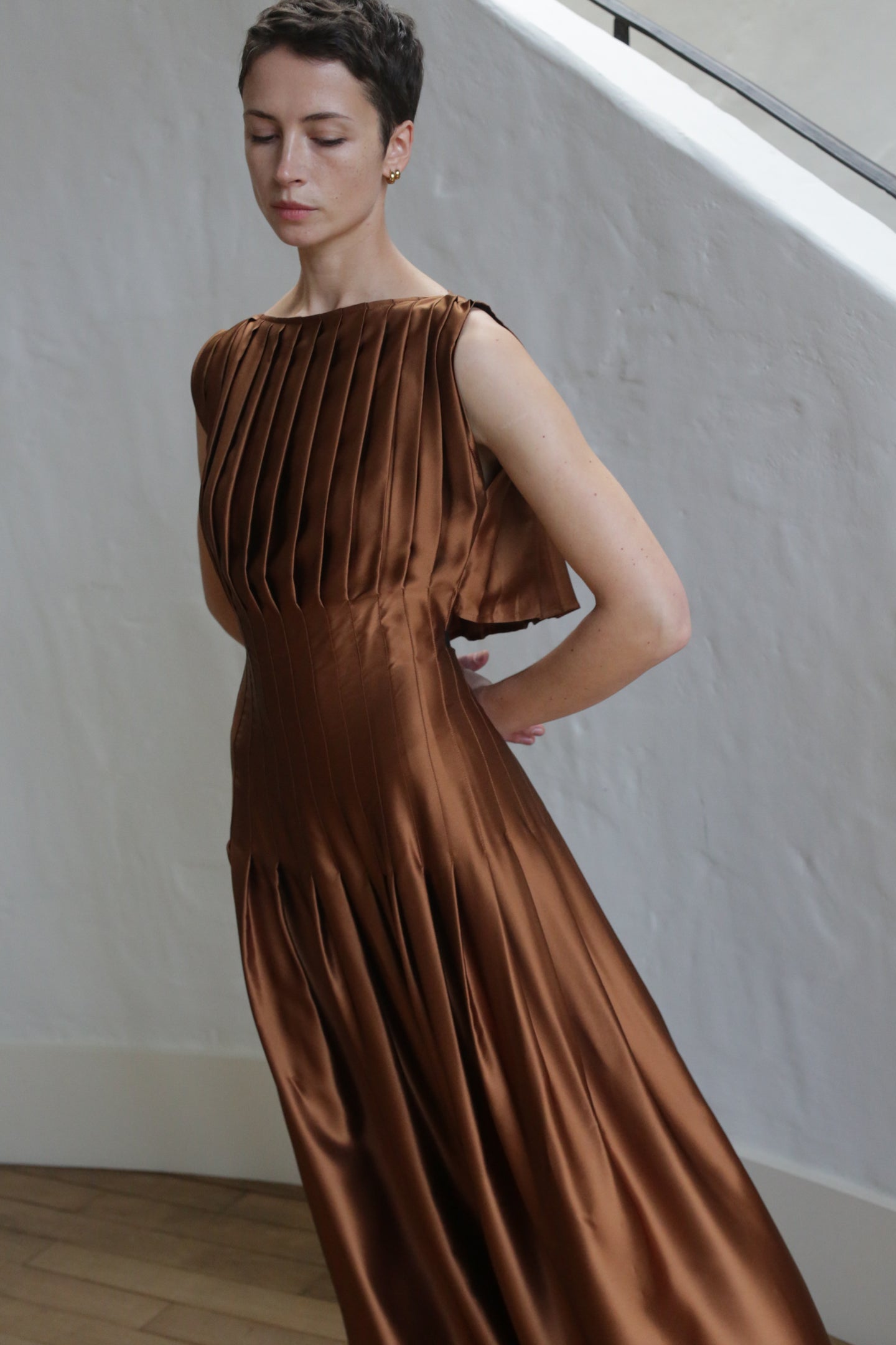 A person with short hair stands indoors, eyes closed, posing in the Hand Pleated Column Dress in copper silk charmeuse. Their arms rest slightly behind their back as they stand against a textured white wall.