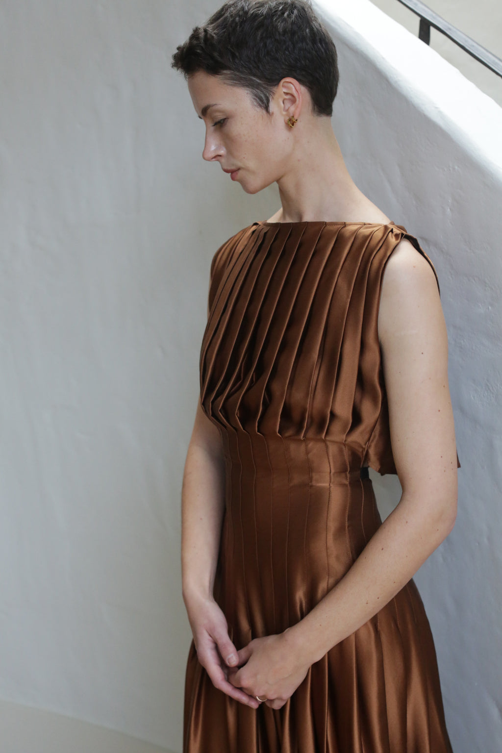 A woman with short dark hair stands indoors by a white wall, looking down with hands clasped, wearing the Hand Pleated Column Dress in copper silk charmeuse.