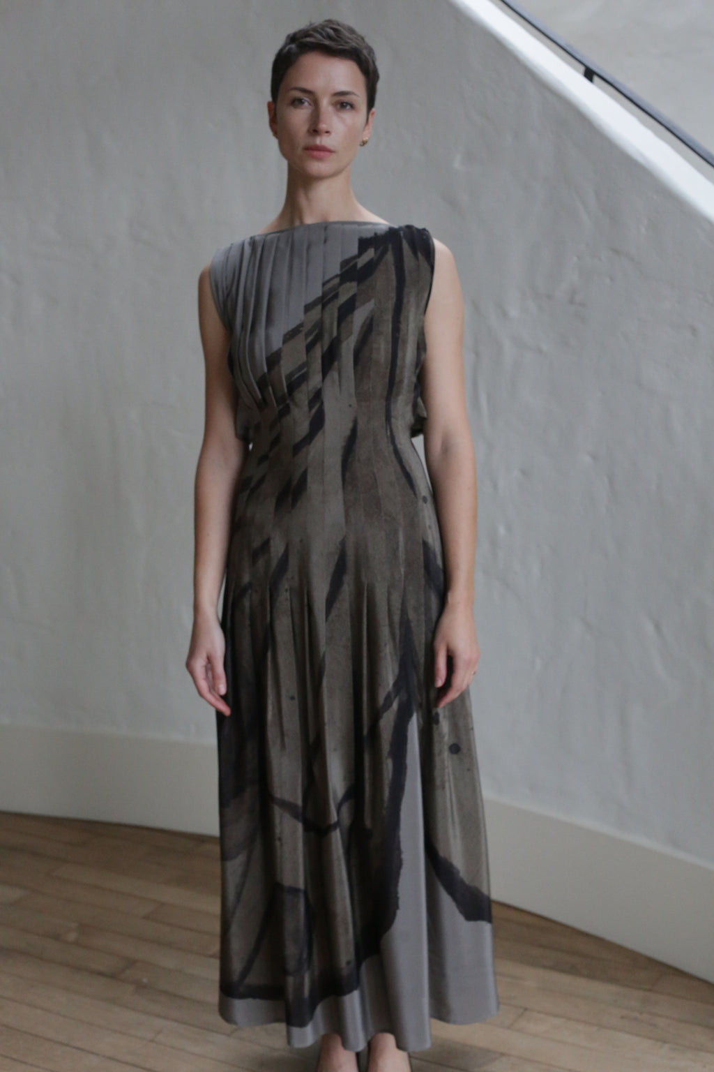 A woman with short hair stands indoors on a wooden floor, wearing the Hand Pleated Column Dress | Butterfly Wing. The sleeveless, long dress features abstract dark patterns. A light-textured wall and window frame are in the background.
