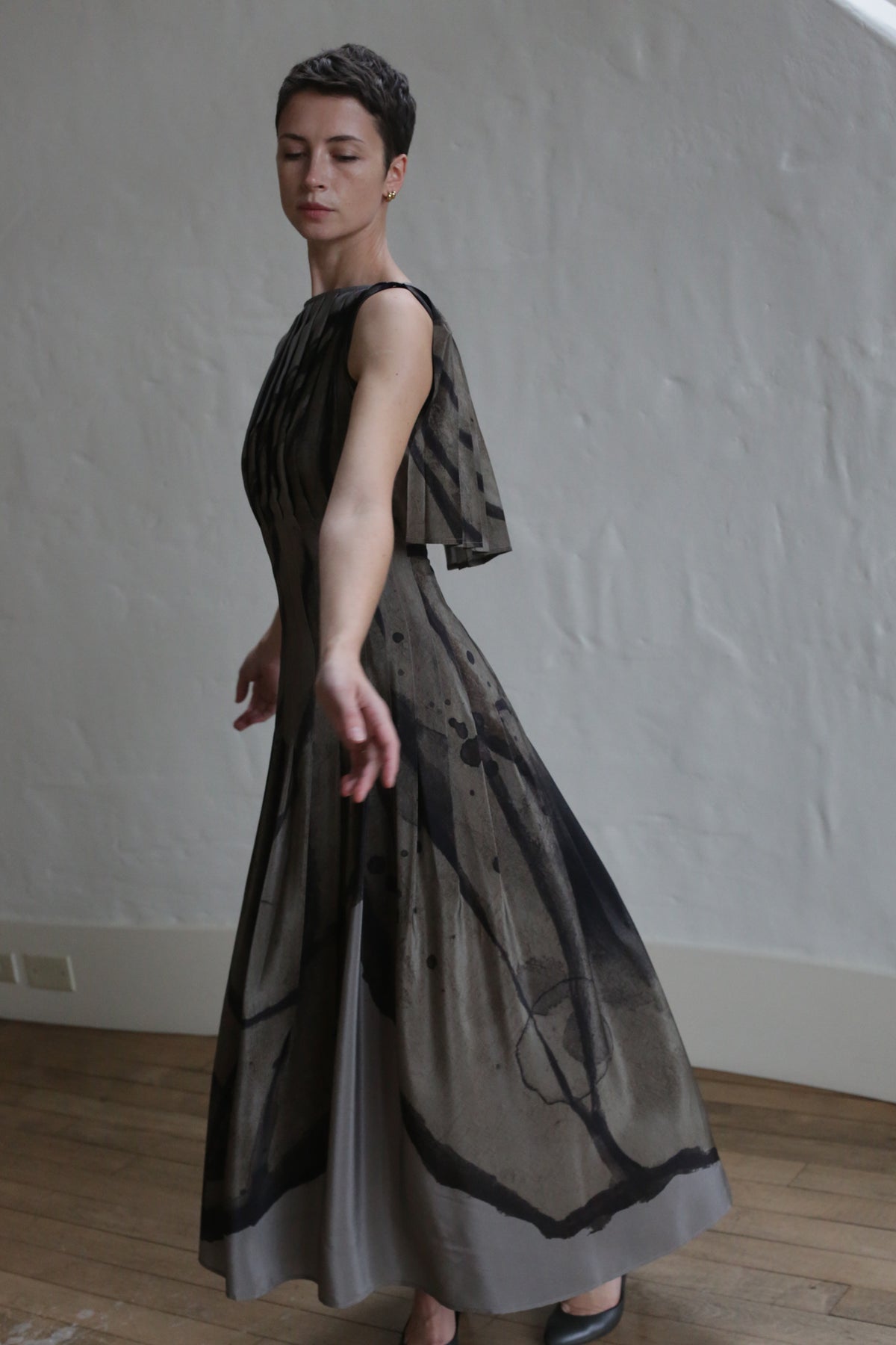 A woman with short hair wears the Hand Pleated Column Dress | Butterfly Wing, a sleeveless flowing dress with abstract black patterns, standing gracefully on a wooden floor before a textured white wall, eyes closed and one arm extended.