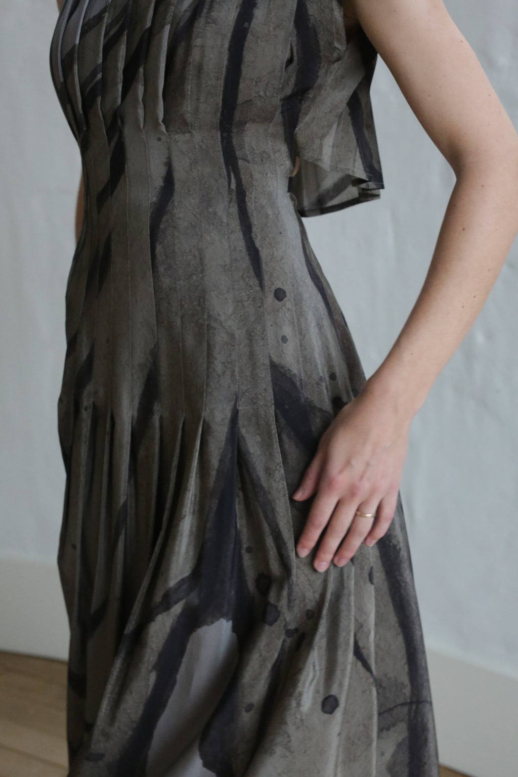 A person wears the Hand Pleated Column Dress | Butterfly Wing, a sleeveless patterned grey and black dress, indoors. Their right hand with a simple ring rests on the skirt; head and lower legs are out of frame.