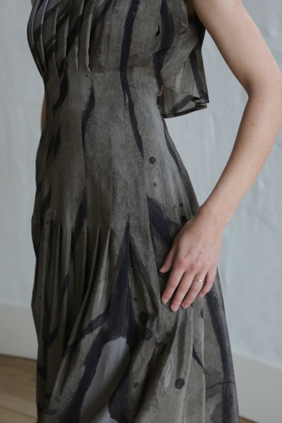A person wears the Hand Pleated Column Dress | Butterfly Wing, a sleeveless patterned grey and black dress, indoors. Their right hand with a simple ring rests on the skirt; head and lower legs are out of frame.