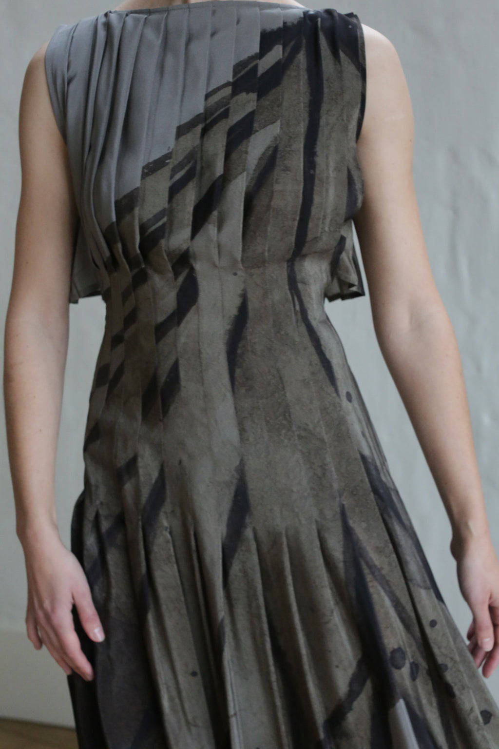 A person models the Hand Pleated Column Dress | Butterfly Wing, a sleeveless dress with geometric black and gray stripes, standing against a plain light background. The head is out of frame.