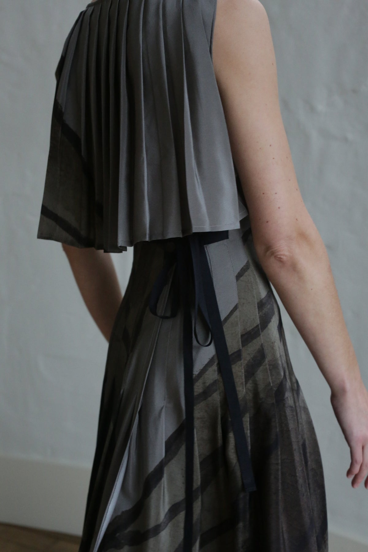 A person is shown from the side and back wearing the Hand Pleated Column Dress | Butterfly Wing, a sleeveless grey dress with dark diagonal stripes and hand-pleated detailing on the upper back, set against a neutral background.
