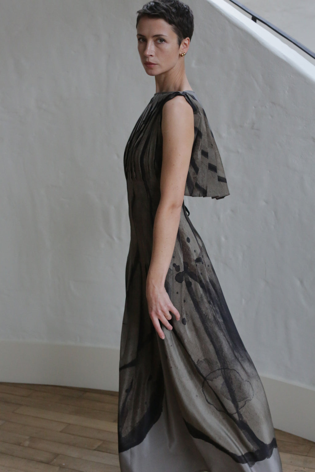 A person with short dark hair stands sideways in front of a white textured wall and wooden floor, wearing the Hand Pleated Column Dress | Butterfly Wing—a long, sleeveless dress with an abstract black and gray pattern.