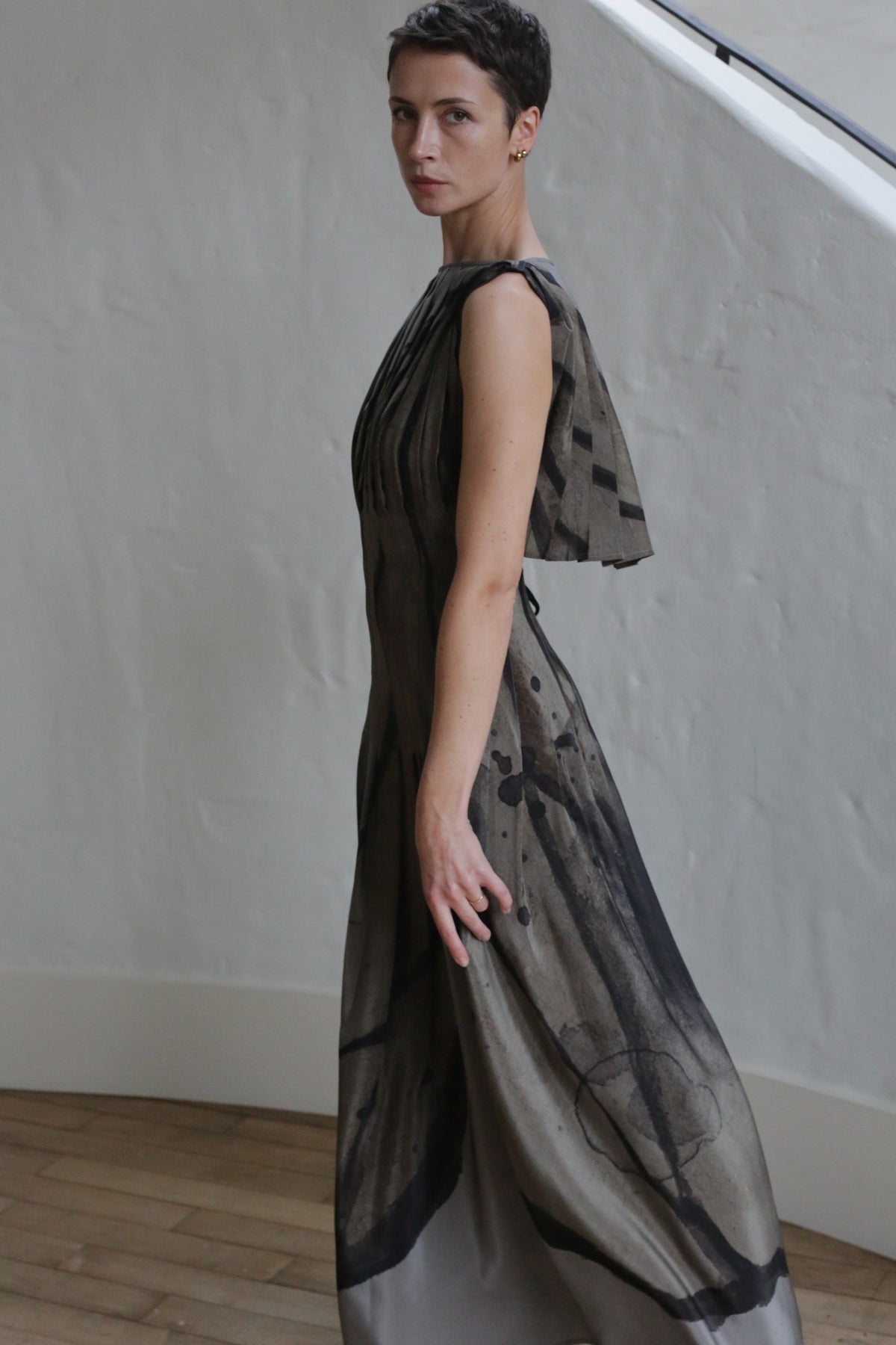 A person with short dark hair stands sideways in front of a white textured wall and wooden floor, wearing the Hand Pleated Column Dress | Butterfly Wing—a long, sleeveless dress with an abstract black and gray pattern.