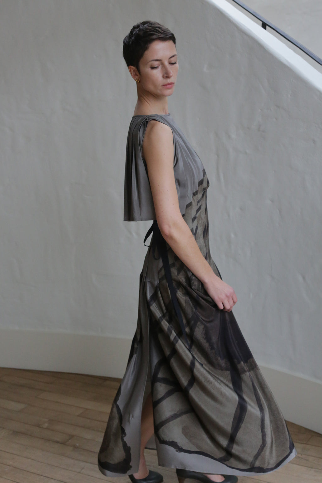 A person with short hair wears the Hand Pleated Column Dress | Butterfly Wing, a flowing gray and black patterned dress, standing indoors on a wooden floor near a white textured wall and staircase, eyes closed with head slightly turned.