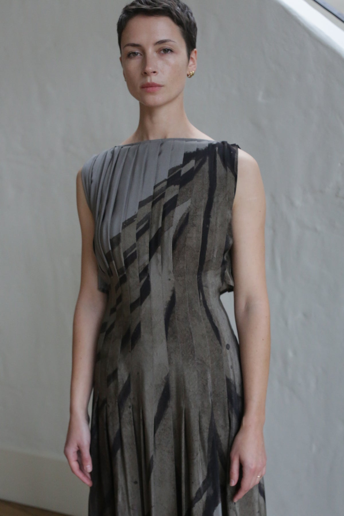 A person with short hair stands indoors against a plain light-colored wall, wearing the Hand Pleated Column Dress | Butterfly Wing—a sleeveless, pleated dress with diagonal black and gray stripes—looking at the camera with a neutral expression.