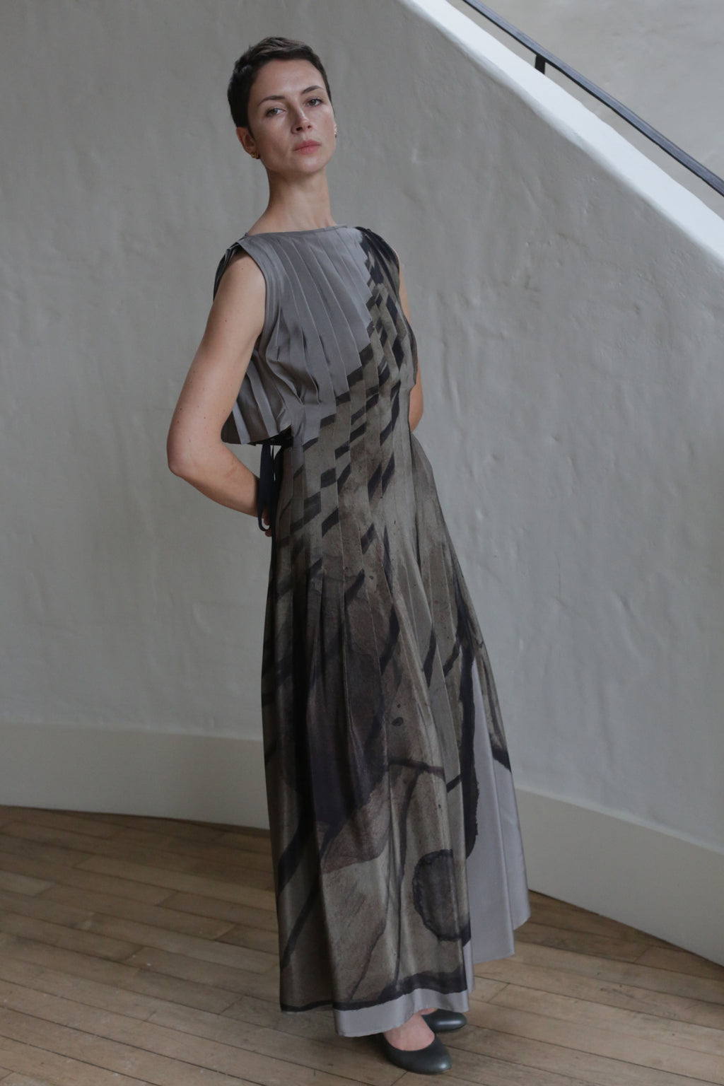 A woman with short hair poses indoors in the Hand Pleated Column Dress | LJ X CL Printed Silk, a floor-length, zero-waste design featuring a geometric abstract print in neutral tones, standing out against wooden floors and a textured white wall.