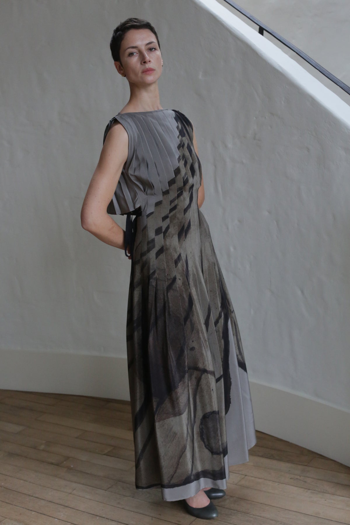A woman with short hair poses indoors in the Hand Pleated Column Dress | LJ X CL Printed Silk, a floor-length, zero-waste design featuring a geometric abstract print in neutral tones, standing out against wooden floors and a textured white wall.