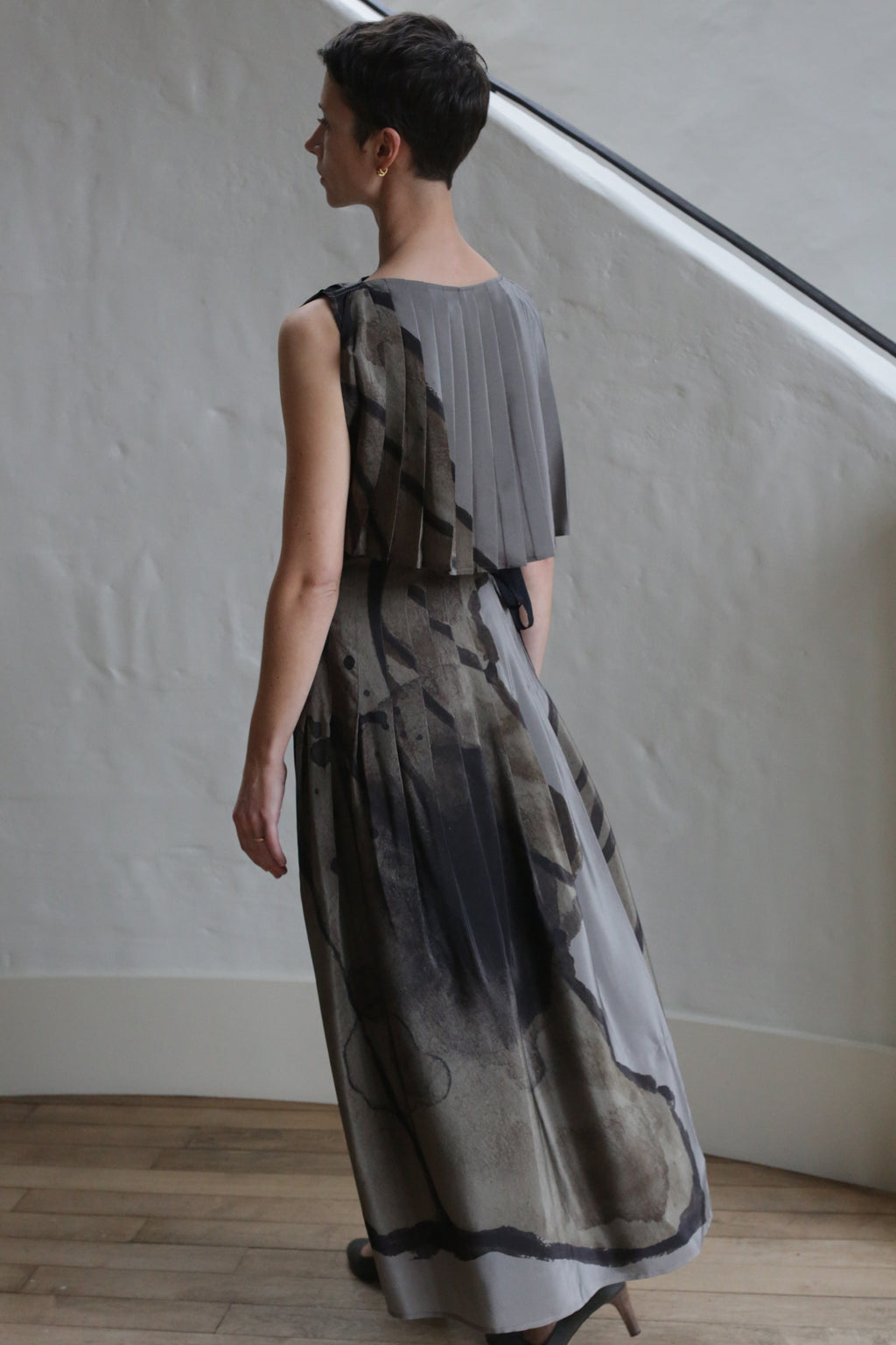 A person with short dark hair wears the Hand Pleated Column Dress | LJ X CL Printed Silk in gray and black, standing indoors on a wooden floor near a white stucco wall and staircase, facing away from the camera.