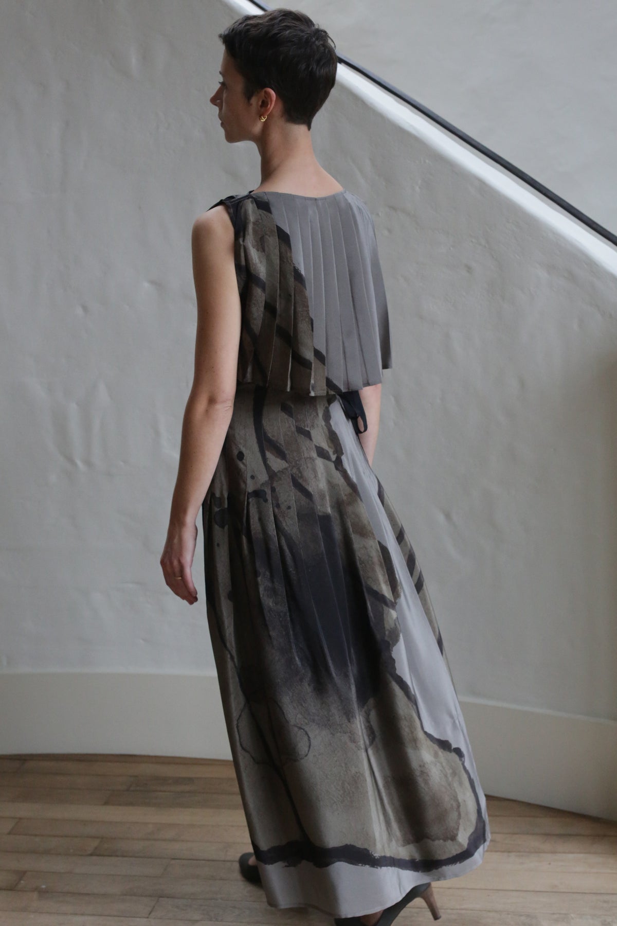 A person with short dark hair wears the Hand Pleated Column Dress | LJ X CL Printed Silk in gray and black, standing indoors on a wooden floor near a white stucco wall and staircase, facing away from the camera.