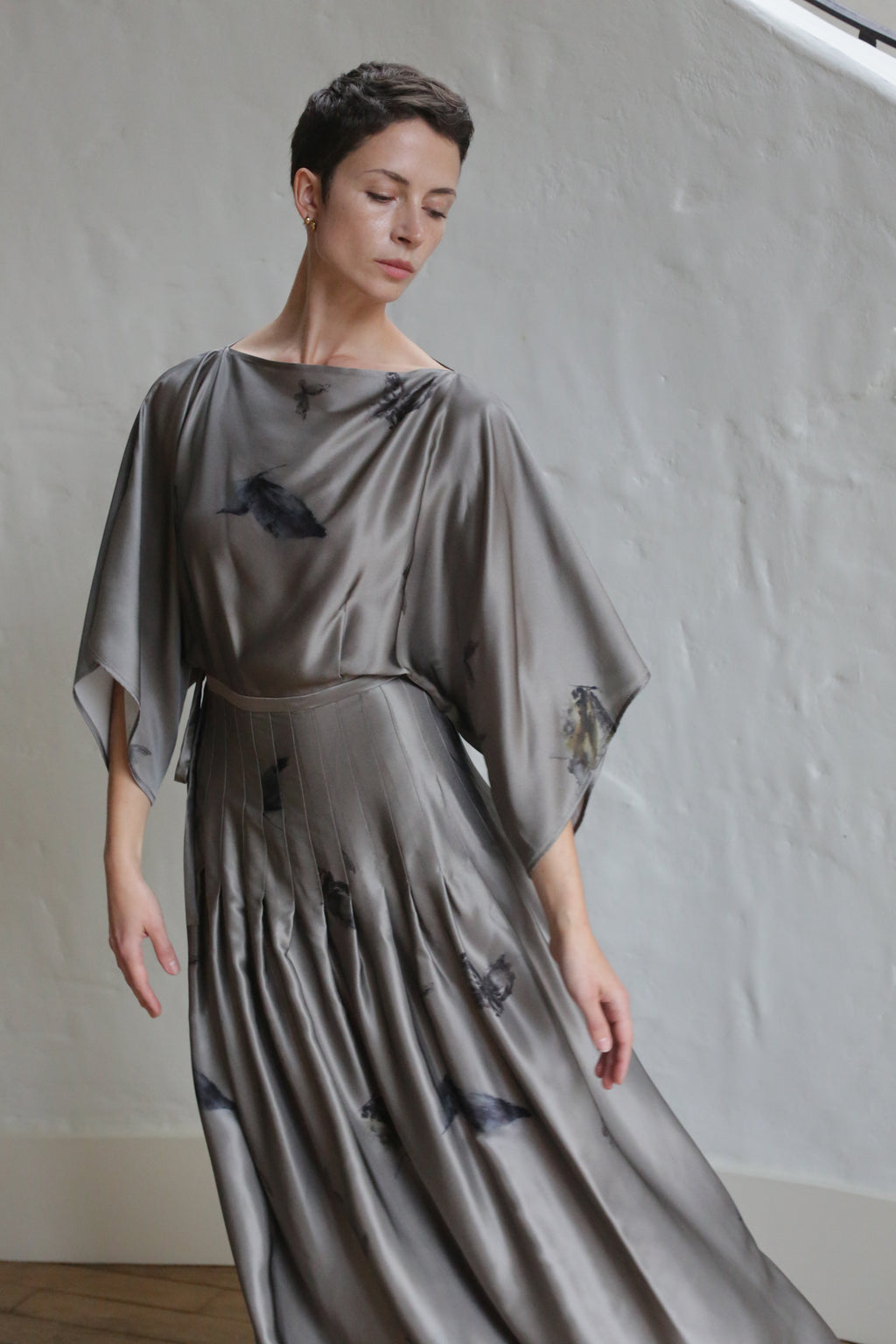 A woman with short hair poses gracefully against a textured white wall, wearing the Hand Pleated Kimono Dress | Small Butterflies—a flowing, silver-gray dress with subtle dark patterns. She looks down, one arm gently extended.