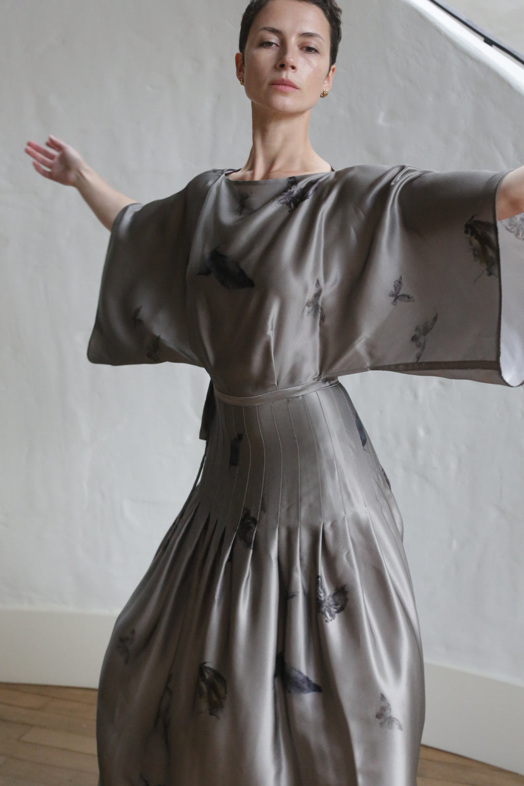 A person with short hair stands indoors against a light wall, arms outstretched, wearing the Hand Pleated Kimono Dress | Small Butterflies—a flowing, shiny grey dress featuring a subtle butterfly pattern.