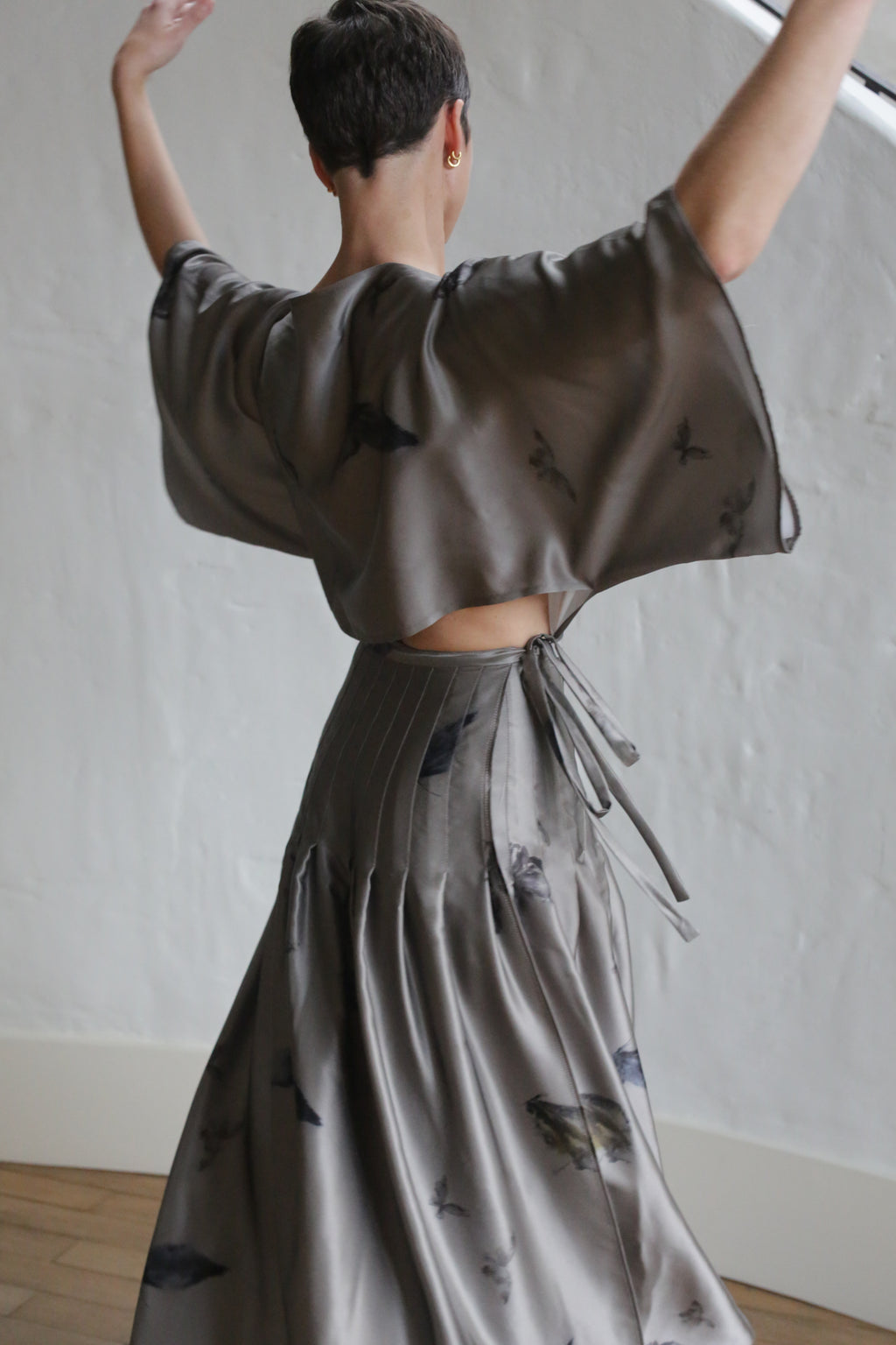 A person with short hair, seen from behind, dances gracefully in the Hand Pleated Kimono Dress | Small Butterflies against a plain white textured wall and light wooden floor. The dress features flowing fabric with wide sleeves and a tie at the waist.