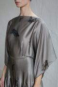 A person wears the Hand Pleated Kimono Dress | Small Butterflies—silky grey with wide sleeves and subtle dark abstract motifs—standing by a light, textured wall, their face partially out of frame.