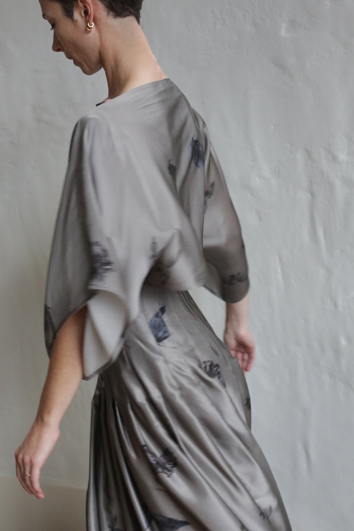 A person in a Hand Pleated Kimono Dress | Small Butterflies, featuring a flowing silhouette and wide sleeves, is pictured mid-motion with their back slightly turned against a plain, light-colored wall.