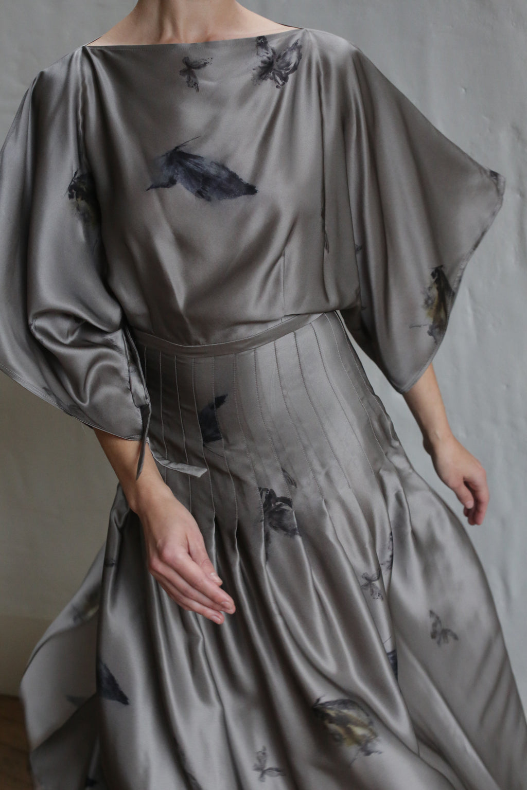 A person wears the Hand Pleated Kimono Dress | LJ X CL Printed Silk—a silver-gray charmeuse kimono dress featuring black feather-like prints, wide sleeves, and pleated details, handmade in San Francisco—against a light textured wall.
