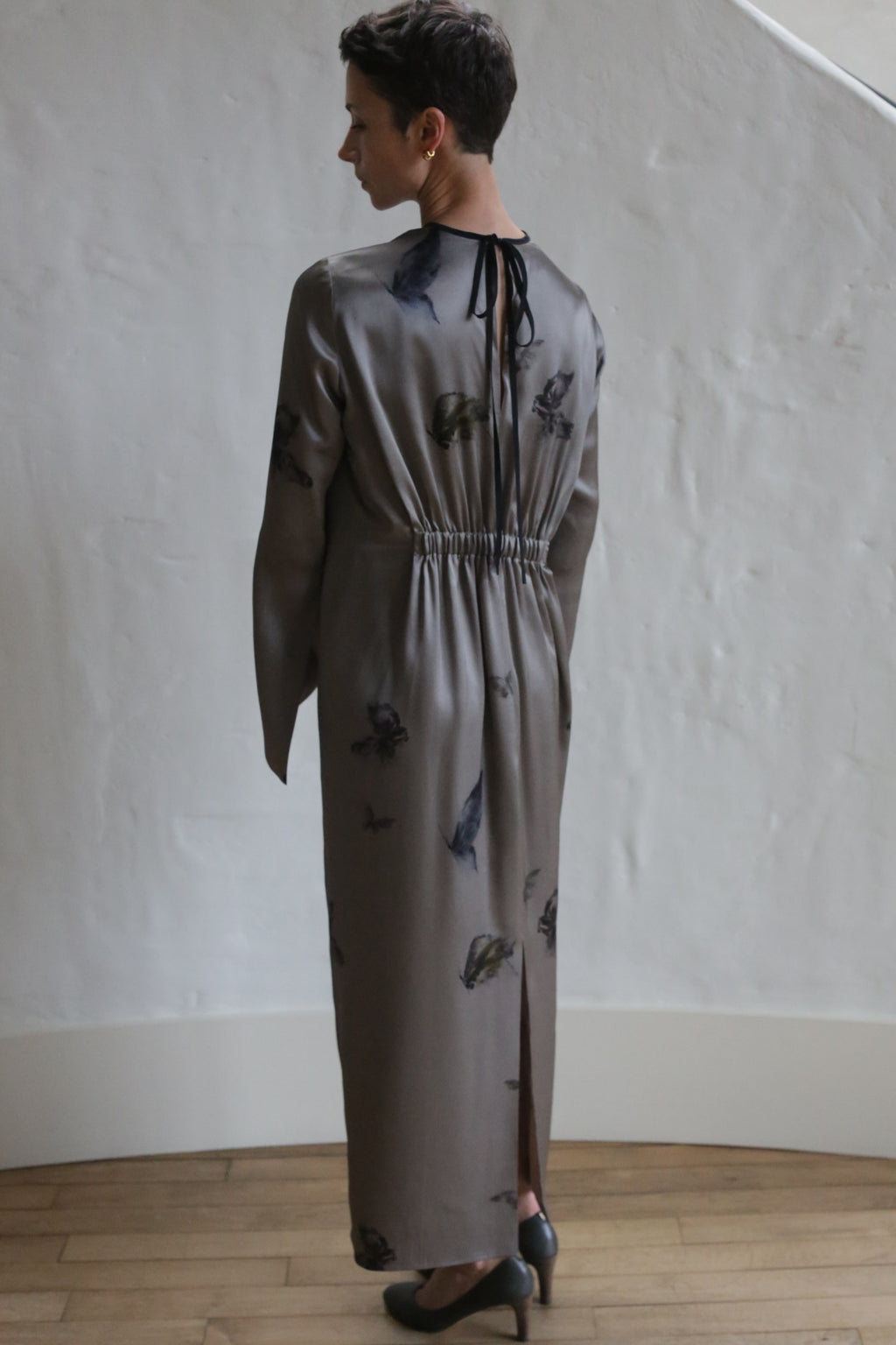 A person with short hair stands indoors, facing a white textured wall and wearing The Butterfly Dress—a long, taupe dress with dark floral patterns, a gathered waist, slit back, neck tie detail, and paired with black heels.