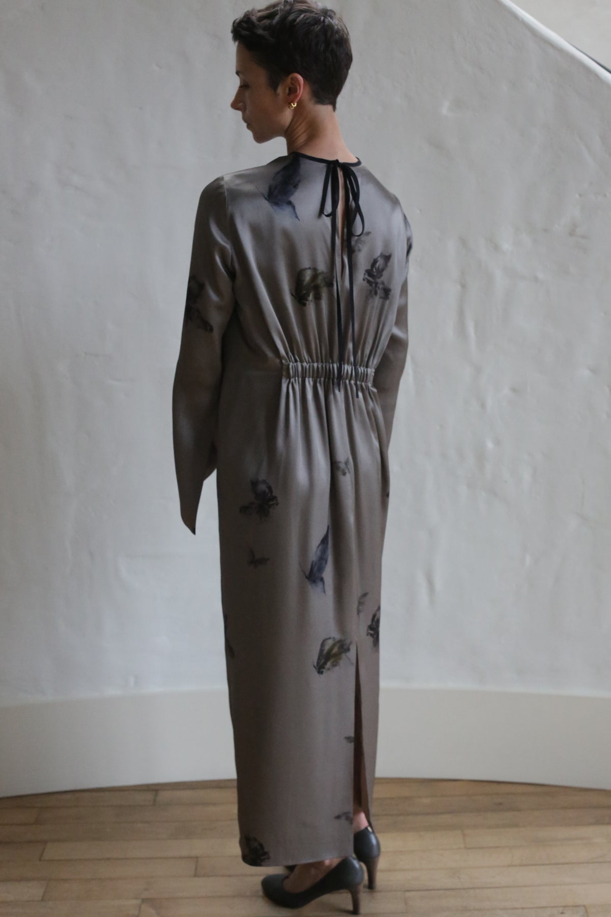 A person with short hair stands indoors, facing a white textured wall and wearing The Butterfly Dress—a long, taupe dress with dark floral patterns, a gathered waist, slit back, neck tie detail, and paired with black heels.
