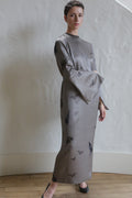 A woman with short hair stands indoors on a wooden floor, wearing The Butterfly Dress—a long, gray kimono-style dress with wide sleeves and a subtle black pattern—posing with one hand on her hip against a textured white wall.