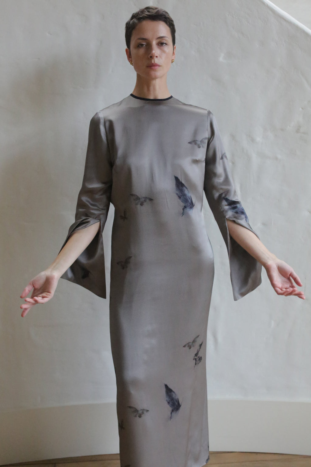 Wearing The Butterfly Dress—a long, silvery-gray gown with subtle dark patterns and open sleeves—a person with short hair stands calmly against a textured, off-white wall, arms gently extended.
