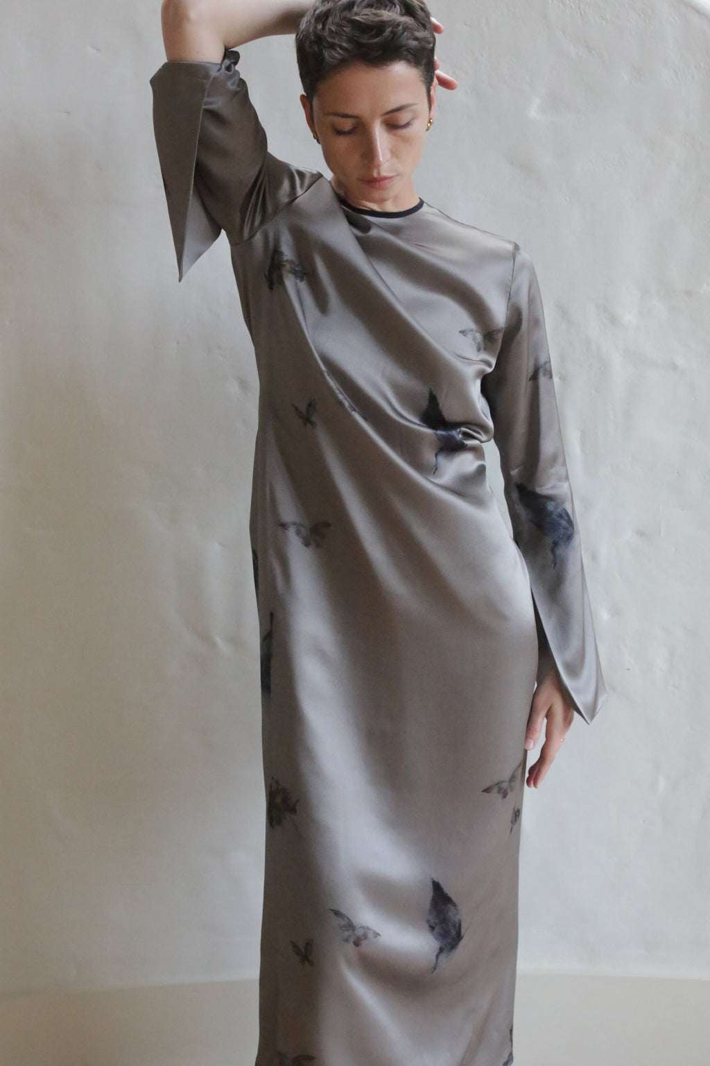 A person with short hair poses in The Butterfly Dress | LJ X CL Printed Silk, a long, loose-fitting handmade silk dress featuring a dark abstract butterfly print, against a plain light background with one arm raised and the other by their side.